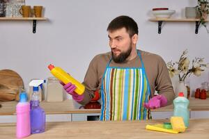 Jeune Beau barbu homme dans le cuisine, nettoie en haut en utilisant détergents, pinceaux, Vaporisateurs photo