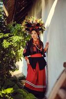 brunette fille dans noir et rouge brodé ukrainien authentique nationale costume et une couronne de fleurs est posant permanent contre une blanc cabane. photo