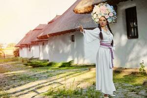 brunette fille dans une blanc ukrainien authentique nationale costume et une couronne de fleurs est posant contre une blanc cabane. photo