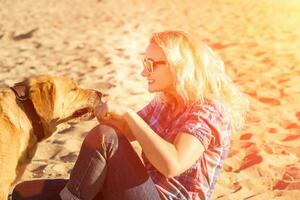 portrait de Jeune magnifique femme dans des lunettes de soleil séance sur le sable plage étreindre d'or retriever chien. fille avec chien par mer. bonheur et amitié. photo