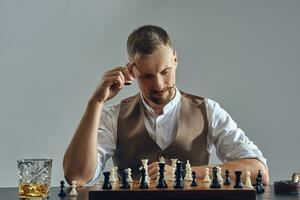 homme avec élégant moustache, habillé dans marron gilet, blanc chemise est en jouant échecs. proche verre de whisky, cendrier, cigare. gris arrière-plan, fermer. photo