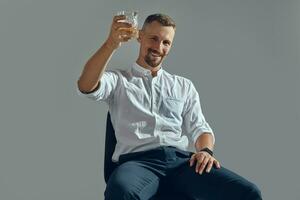 homme avec élégant moustache, dans classique blanc chemise et bleu pantalon est séance sur chaise, en portant verre de whisky. gris arrière-plan, fermer tir. photo