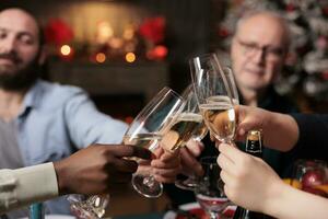 joyeux les personnes applaudissement avec de l'alcool ensemble à dîner table pendant Noël veille festivité, famille tintement du vin des lunettes à maison. diverse gens profiter décembre hiver vacances tradition. photo