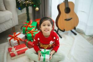 Noël, Noël, hiver concept. asiatique bébé enfant fille en portant cadeau des boites souriant content joyeux. fête le content Nouveau année près le Noël arbre dans une vivant pièce à maison. photo