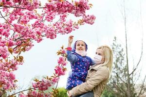 une européen mère dans une respirateur avec sa fille sont permanent près floraison arbre. protecteur masque à enregistrer se de virus. photo