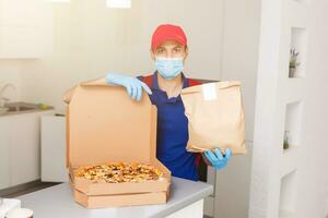 livraison homme employé dans rouge casquette T-shirt uniforme masque gants donner nourriture commande Pizza des boites isolé sur Jaune Contexte studio. un service quarantaine pandémie coronavirus virus grippe 2019-ncov concept photo