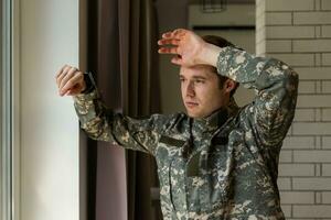 sérieux cheveux courts Jeune homme dans militaire uniforme soldat à la recherche par le fenêtre, dérangé soldat Souffrance de post-traumatique syndrome après de retour maison, copie espace, fermer. photo