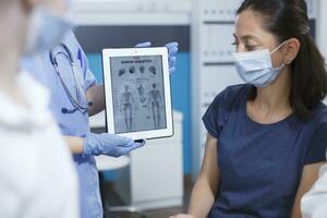 fermer coup de une la personne en portant tablette dans clinique Bureau à Explique médical se soucier à femelle patient. sélectif concentrer de une Dame portant visage masque et à la recherche à le Humain squelette image sur appareil. photo