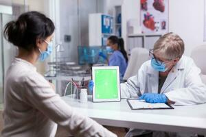 personnes âgées médecin en portant tablette avec vert écran dans clinique pièce Parlant avec Jeune patient expliquant le traitement à la recherche à maquette afficher. montrant à isoler moniteur, chrominance clé bureau. photo