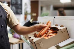 Commerçant remplit en haut caisses sur faible carbone empreinte zéro déchets magasin étagères avec localement grandi des légumes. magasinier réapprovisionnements local les courses boutique avec nourriture articles, proche en haut coup sur carottes photo