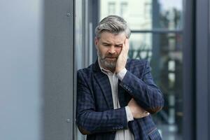 mature patron malade, homme a sévère mal de tête, homme d'affaire dans affaires costume adulte surmené en portant mains derrière diriger, gris aux cheveux ouvrier à l'extérieur Bureau bâtiment avec mal de tête. photo