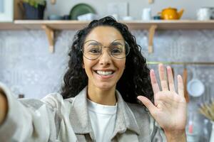 femme avec frisé cheveux et des lunettes à la recherche dans téléphone intelligent caméra prise selfie et parlant sur vidéo appel à maison, hispanique femme souriant à Accueil dans cuisine, agitant salutation geste. photo