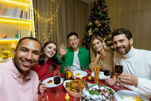 une entreprise de interracial Jeune gens séance à une de fête Nouveau années table et prise une selfie ensemble sur une mobile téléphone. elles ou ils tenir des lunettes dans leur mains, vague à le caméra, sourire, réjouir. photo