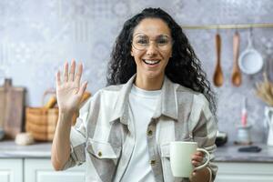 Jeune magnifique hispanique femme parlant et agitant à le caméra pendant une vidéo appel. permanent dans le cuisine à maison, en portant une tasse, souriant, court le sien Blog. photo