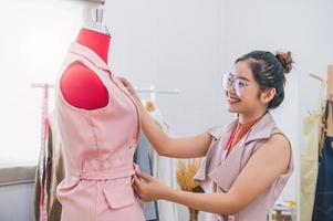 Happy asian female fashion designer girl faisant s'adapter sur les vêtements d'uniforme de robe formelle sur le modèle de mannequin. showroom élégant de créateur de mode. concept de couture et de tailleur. styliste couturière créative photo