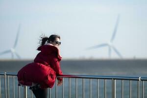 sur le Contexte de Moulins à vent, une Jeune femme dans une rouge veste est profiter sa hiver vacances. photo