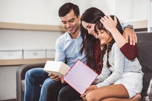 parents de famille heureux et petite fille regardant dans une boîte-cadeau à noël et le jour de l'an sur un canapé dans le salon. cadeau de Noël pour les bons enfants surpris dans une maison heureuse. concept de personnes et de modes de vie photo