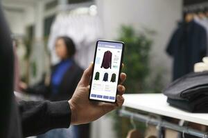 caucasien client dans une vêtements boutique est en portant une téléphone intelligent et à la recherche à vêtements sur une boutique site Internet. Masculin Shopaholic est lire attentivement Vêtements articles dans une centre commercial en utilisant une téléphone portable. photo