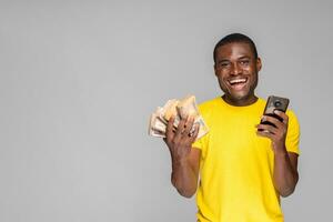 excité Jeune africain homme en portant en espèces et en utilisant le sien téléphone réjouissance photo