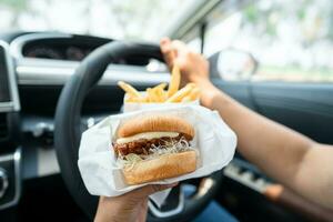 asiatique Dame en portant Hamburger et français frites à manger dans voiture, dangereux et risque un accident. photo