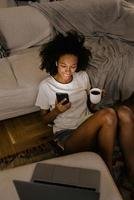 jeune femme noire dans des écouteurs utilisant un téléphone portable et buvant du café