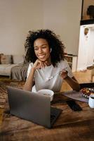 jeune femme noire dans les écouteurs à l'aide d'un ordinateur portable et prenant son petit déjeuner