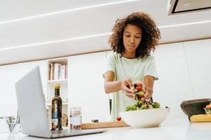 jeune femme noire faisant de la salade tout en utilisant un ordinateur portable dans la cuisine photo