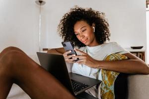 jeune femme noire utilisant un téléphone portable et un ordinateur portable tout en se reposant sur un canapé