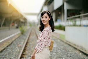 Jeune asiatique femme voyageur avec tissage panier content souriant à la recherche à une caméra à côté de train chemin de fer. périple voyage mode de vie, monde Voyage explorateur ou Asie été tourisme concept. photo