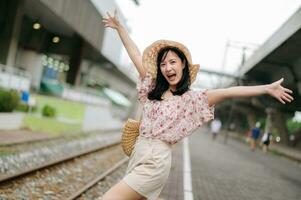 Jeune asiatique femme voyageur avec tissage panier content souriant à la recherche à une caméra à côté de train chemin de fer. périple voyage mode de vie, monde Voyage explorateur ou Asie été tourisme concept. photo