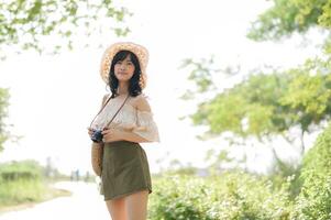 portrait de asiatique Jeune femme voyageur avec tissage chapeau et panier et une caméra sur vert Publique parc la nature Contexte. périple voyage mode de vie, monde Voyage explorateur ou Asie été tourisme concept. photo