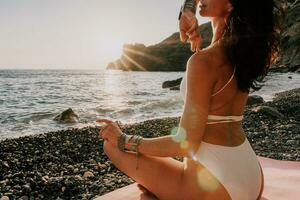 femme mer yoga. content femme dans blanc maillot de bain et boho style bracelets pratiquant en plein air sur yoga tapis par mer sur le coucher du soleil. femmes yoga aptitude routine. en bonne santé mode de vie, harmonie et méditation photo