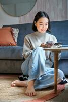 La technologie et personnes. Jeune élégant asiatique femme est assis à Accueil avec sa téléphone intelligent, envoyer des SMS message, en utilisant application sur mobile téléphone photo