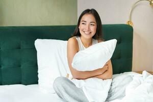 romantique souriant asiatique fille mensonge dans sa lit, étreindre oreiller et regarder rêveur, paresseux les matins dans chambre photo