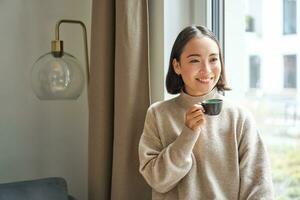 portrait de magnifique asiatique femme séance à Accueil avec tasse de café, profiter sa Expresso tandis que à la recherche à l'extérieur fenêtre à passant, souriant Heureusement photo