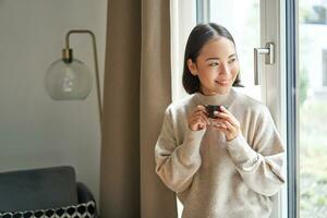 portrait de magnifique asiatique femme séance à Accueil avec tasse de café, profiter sa Expresso tandis que à la recherche à l'extérieur fenêtre à passant, souriant Heureusement photo