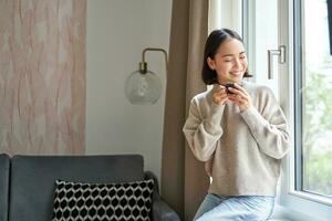 portrait de Jeune asiatique femme séance près fenêtre et à la recherche dehors, en buvant chaud café de Expresso tasse et profiter sa confortable journée de à Accueil photo