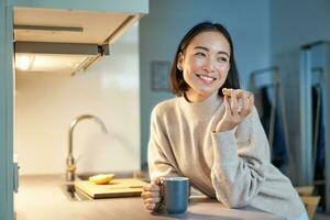 magnifique Jeune asiatique femme en mangeant pain grillé sur sa cuisine, en buvant café et souriant photo