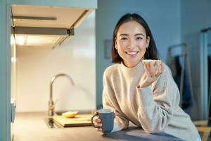 magnifique Jeune asiatique femme en mangeant pain grillé sur sa cuisine, en buvant café et souriant photo