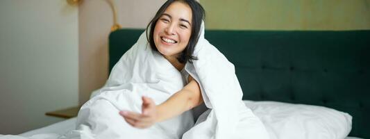 magnifique asiatique femme séance sur lit, couvert avec blanc couette, souriant, profiter content fin de semaine matin, en riant à caméra photo