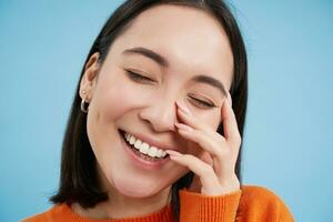 proche en haut portrait de Candide content femme, asiatique fille en riant et souriant, touche sa clair peau, spectacles blanc dents, bleu Contexte photo