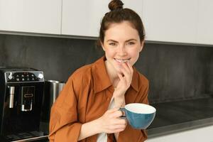 portrait de magnifique Jeune femme en buvant café, permanent dans le cuisine avec gros tasse, profiter Frais aromatique haricots, délicieux cappuccino photo