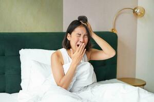 femme veille en haut dans lit, bâillement et grimaçant, de bonne heure Matin après dormir, séance dans chambre photo