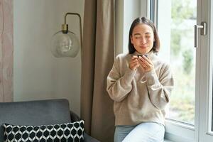 magnifique Jeune asiatique femme séance près fenêtre et en buvant sa café, en portant Expresso tasse et à la recherche à l'extérieur avec détendu, souriant visage expression photo