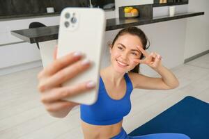 portrait de fille prend selfie sur téléphone intelligent tandis que Est-ce que sa faire des exercices entraînement. Jeune femme Est-ce que sa des exercices à maison, prend selfie pour des sports Blog, est assis dans vivant pièce photo