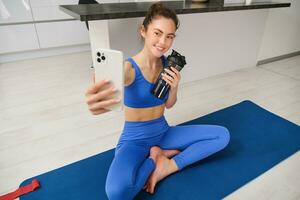 portrait de femme prise selfie avec l'eau bouteille, aptitude instructeur spectacles sa des exercices, Faire faire des exercices de maison, sur caoutchouc bleu yoga tapis, portant tenue de sport photo