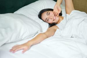 romantique asiatique fille veille en haut dans confortable chambre à coucher, mensonge dans lit avec blanc feuilles, élongation sa main vers vide oreiller photo