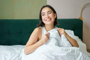 proche en haut de content magnifique asiatique femme, veille en haut dans lit et jouit matin, à la recherche à l'extérieur fenêtre avec somnolent sourire sur sa affronter, étreindre couette photo