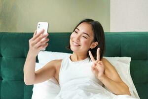 magnifique asiatique fille mensonge dans lit, fabrication Matin selfie, prise image sur téléphone intelligent dans chambre à coucher, souriant Heureusement photo