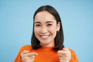 je t'ai eu. proche en haut portrait de souriant asiatique femme points les doigts à caméra heureux, félicite toi, des stands plus de bleu Contexte photo